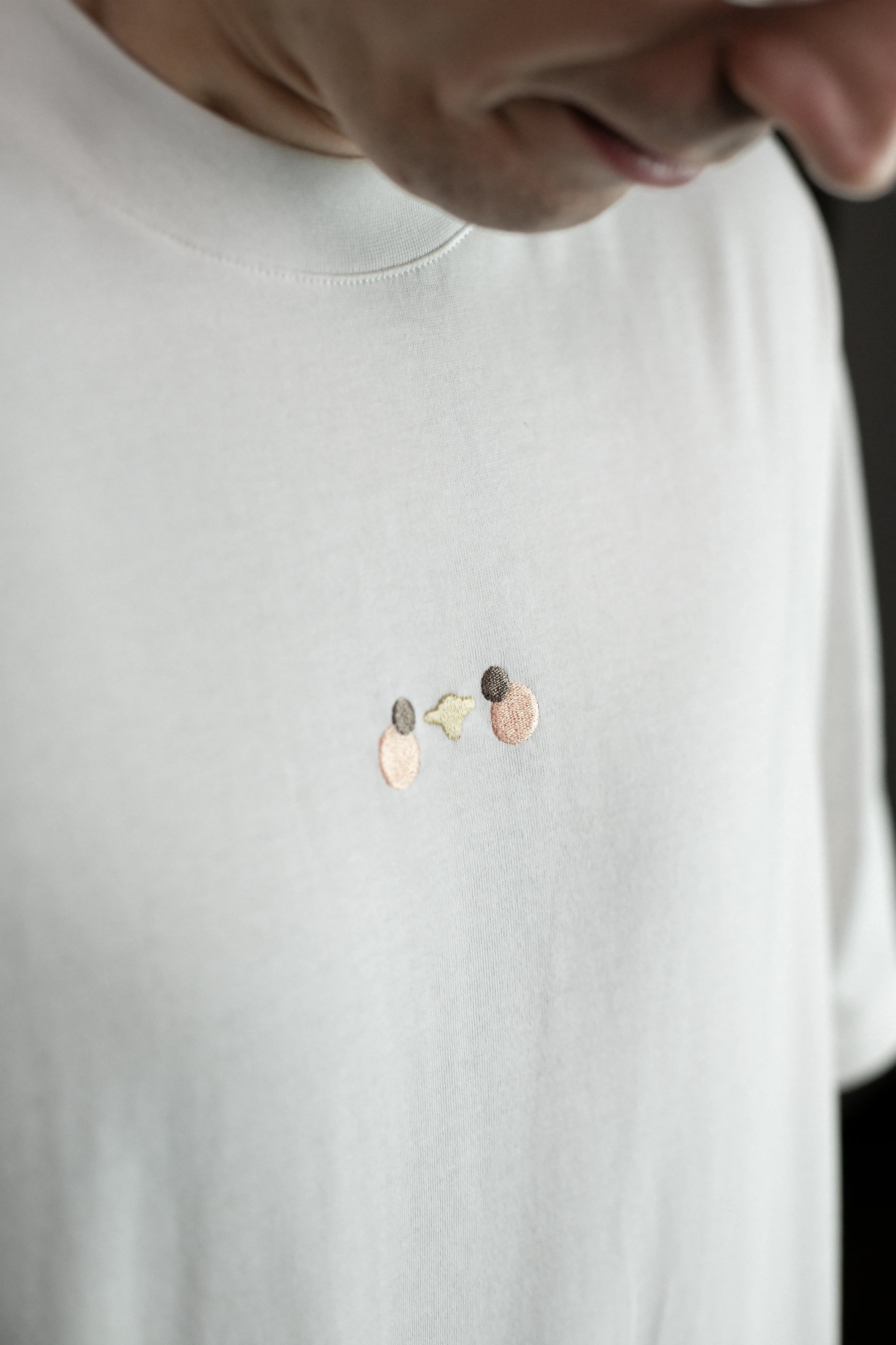 Detailansicht der Vorderseite des weißen T-Shirts mit kleiner, minimalistischer Stickerei in der Mitte der Brust. Die Stickerei zeigt drei kleine Kreise in Beige, Grau und Gelb – eine abstrahierte Darstellung der Möwe aus der Kollektion.