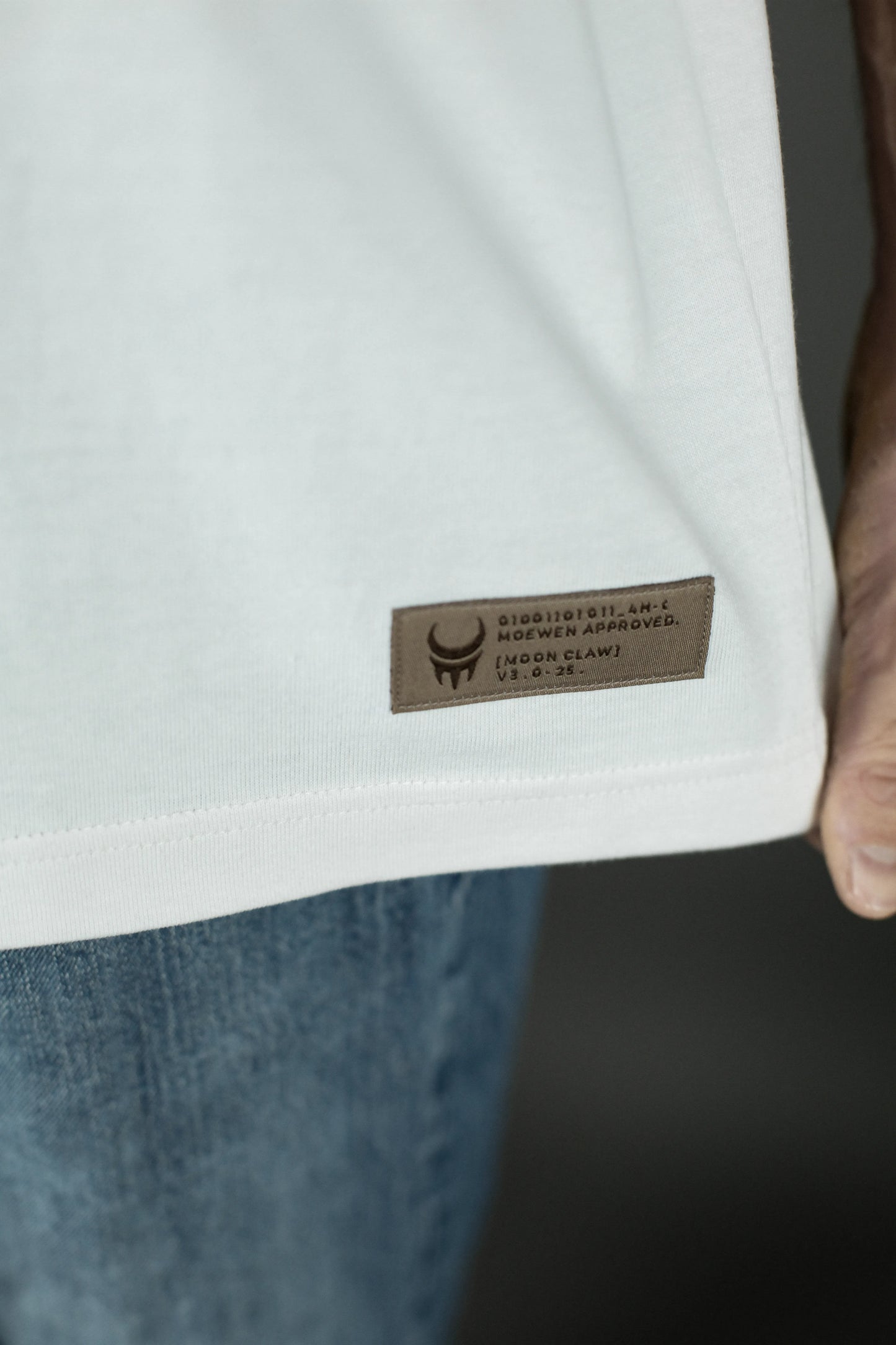 Nahaufnahme des unteren Saums des weißen T-Shirts mit rechteckigem, braunem Label. Darauf steht „MOEWEN APPROVED – MOONCLAW V3.0.25“ mit einem kleinen Symbol in Schwarz.
