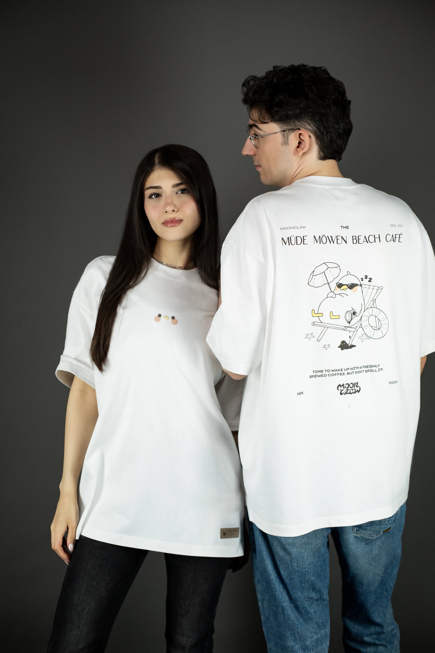 Zwei Personen tragen das weiße T-Shirt: Eine zeigt die Vorderseite mit der kleinen Stickerei auf der Brust, die andere steht mit dem Rücken zur Kamera und zeigt den großflächigen Rückendruck „MÜDE MÖWEN BEACH CAFÉ“ mit Möwenmotiv.