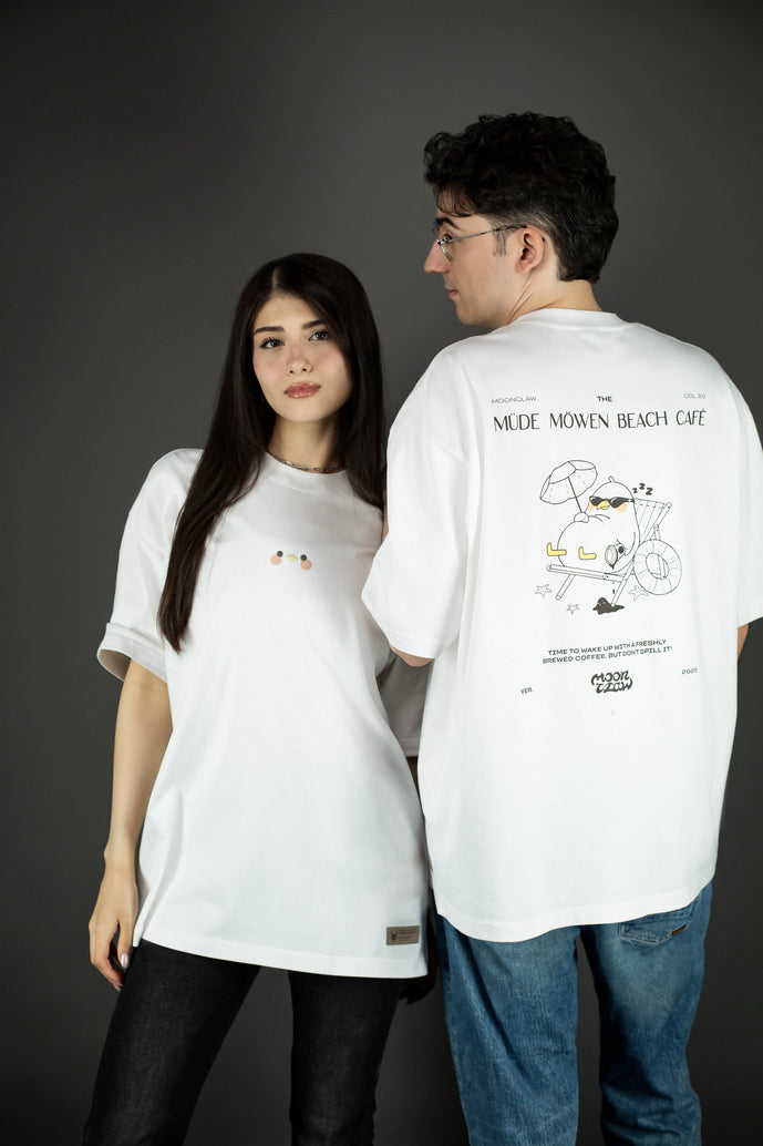 Zwei Personen tragen das weiße T-Shirt: Eine zeigt die Vorderseite mit der kleinen Stickerei auf der Brust, die andere steht mit dem Rücken zur Kamera und zeigt den großflächigen Rückendruck „MÜDE MÖWEN BEACH CAFÉ“ mit Möwenmotiv.