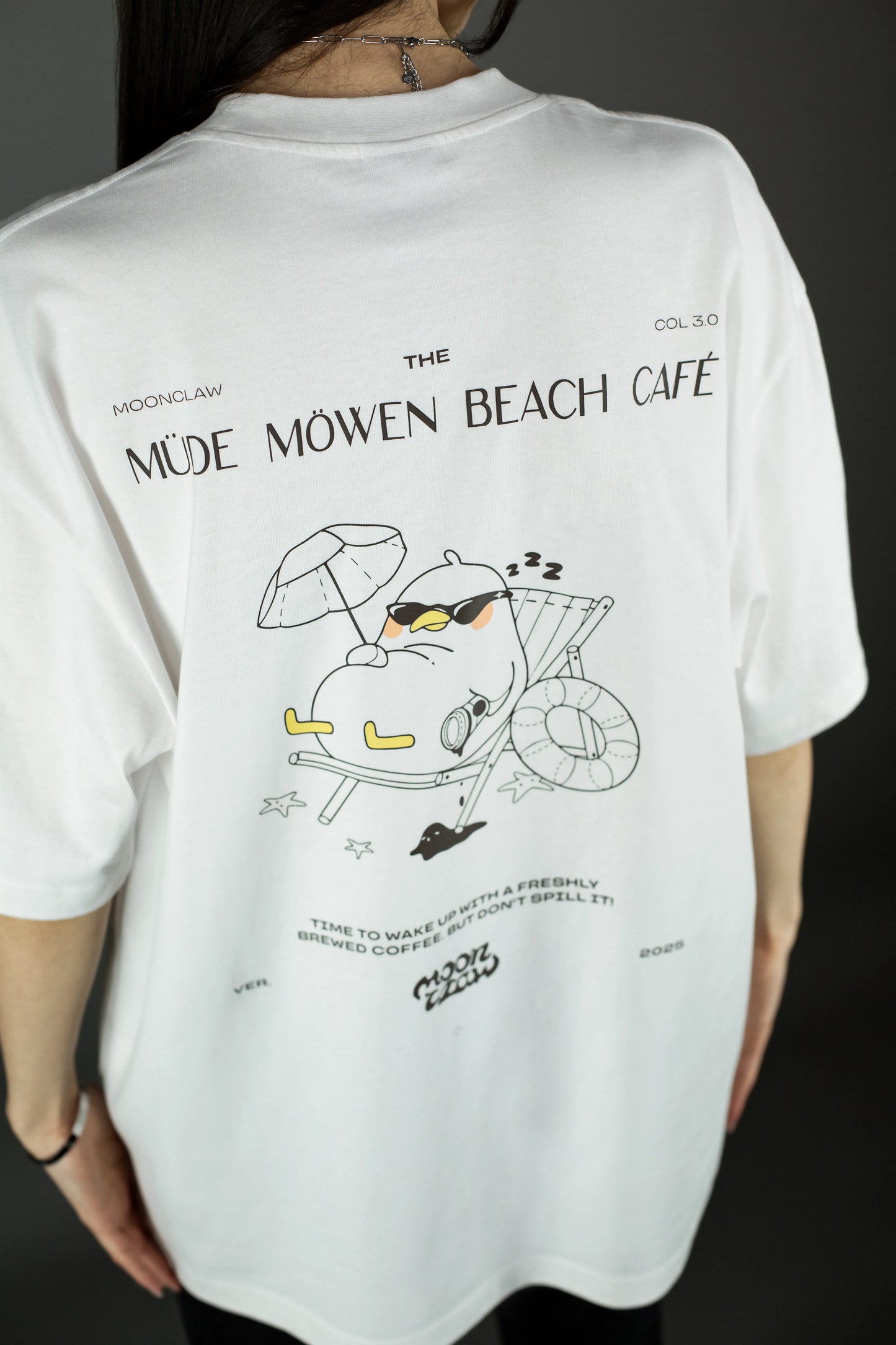 Rückansicht einer Person mit weißem T-Shirt. Der großflächige Rückendruck zeigt die schlafende Möwe mit Sonnenbrille und Kaffeebecher im Liegestuhl unter einem Sonnenschirm, ergänzt durch den Schriftzug „MÜDE MÖWEN BEACH CAFÉ“.