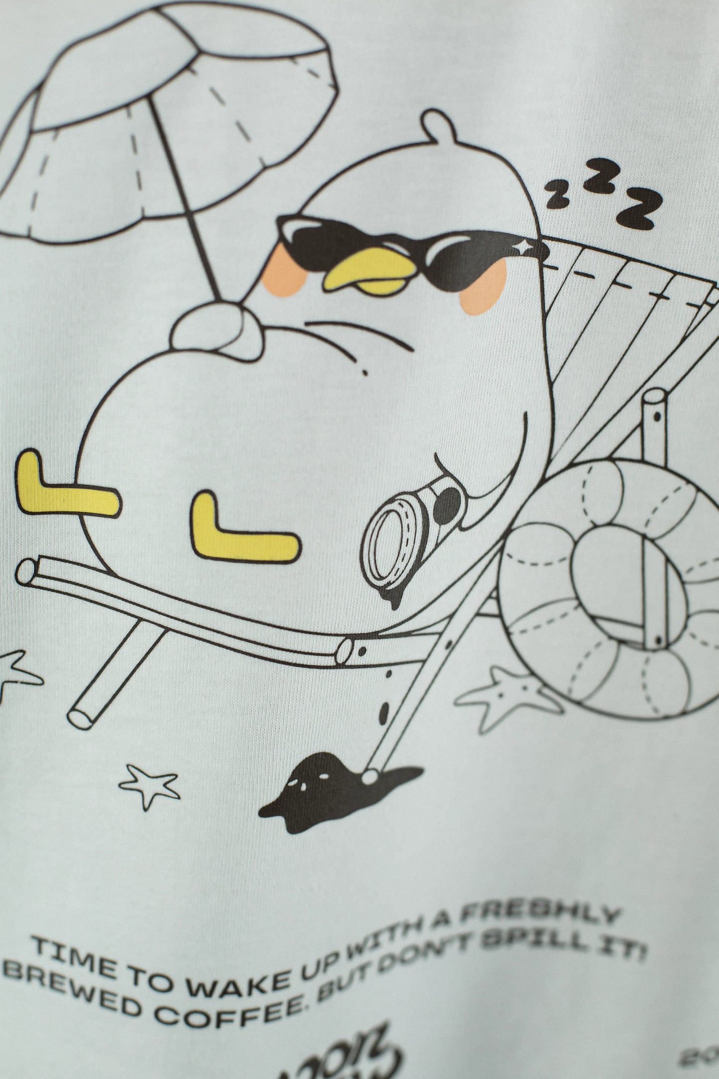 Nahaufnahme des Rückendrucks auf einem weißen T-Shirt. Das Motiv zeigt eine entspannte Möwe mit Sonnenbrille, die auf einem Liegestuhl unter einem Sonnenschirm sitzt. Neben ihr liegen ein Rettungsring, eine Kaffeetasse und kleine Seesterne. Darunter steht der Text „Time to wake up with a freshly brewed coffee. But don’t spill it!“.