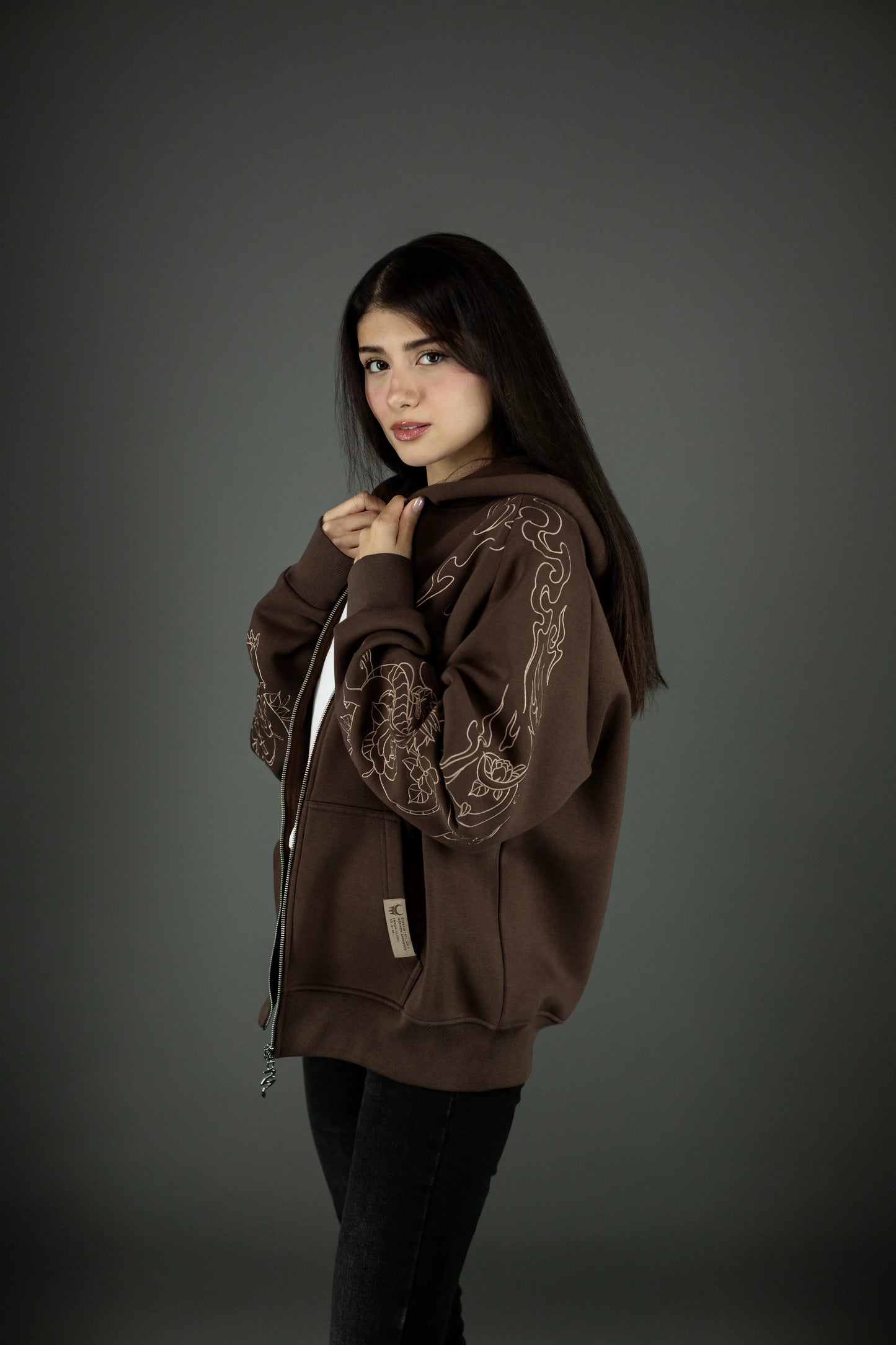 Seitenansicht des braunen Hoodies mit beigem Drachenmuster entlang des Ärmels. Das Motiv zeigt einen stilisierten Drachen mit Blumen und Flammenlinien. Die Kapuze liegt locker auf dem Rücken.
