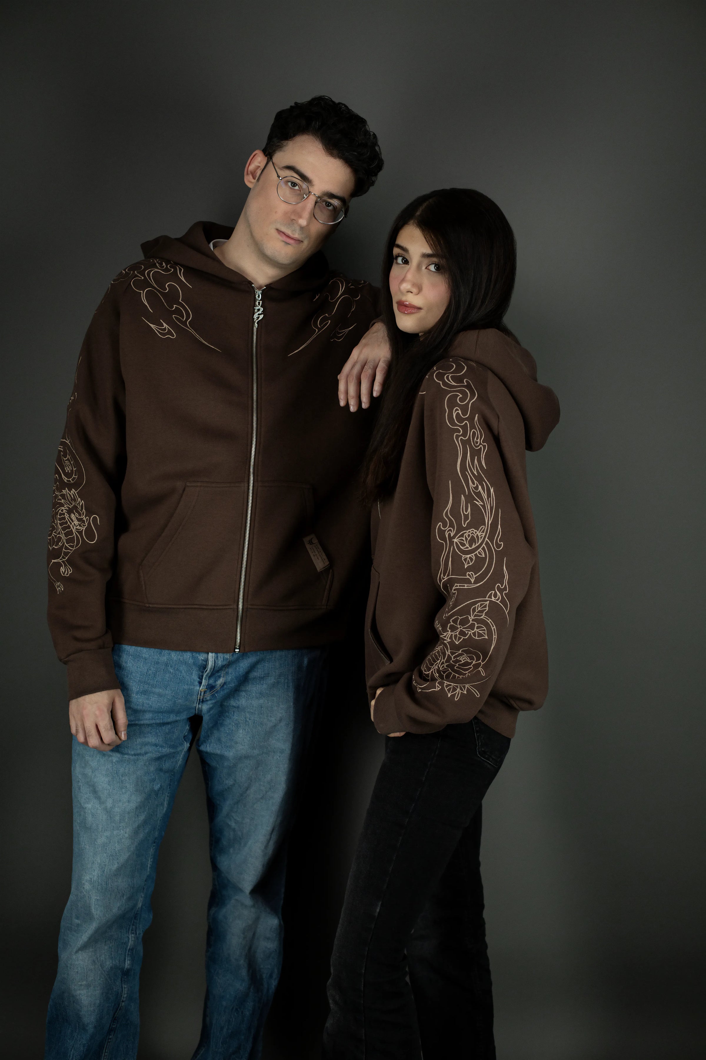 Zwei Personen tragen einen braunen Zip-Hoodie mit beigem Drachenmotiv auf den Ärmeln. Der Hoodie zeigt vorn eine Kängurutasche und Metallreißverschluss. Das Motiv läuft von der Schulter über den Arm und kombiniert Flammen- mit Blumenornamenten.