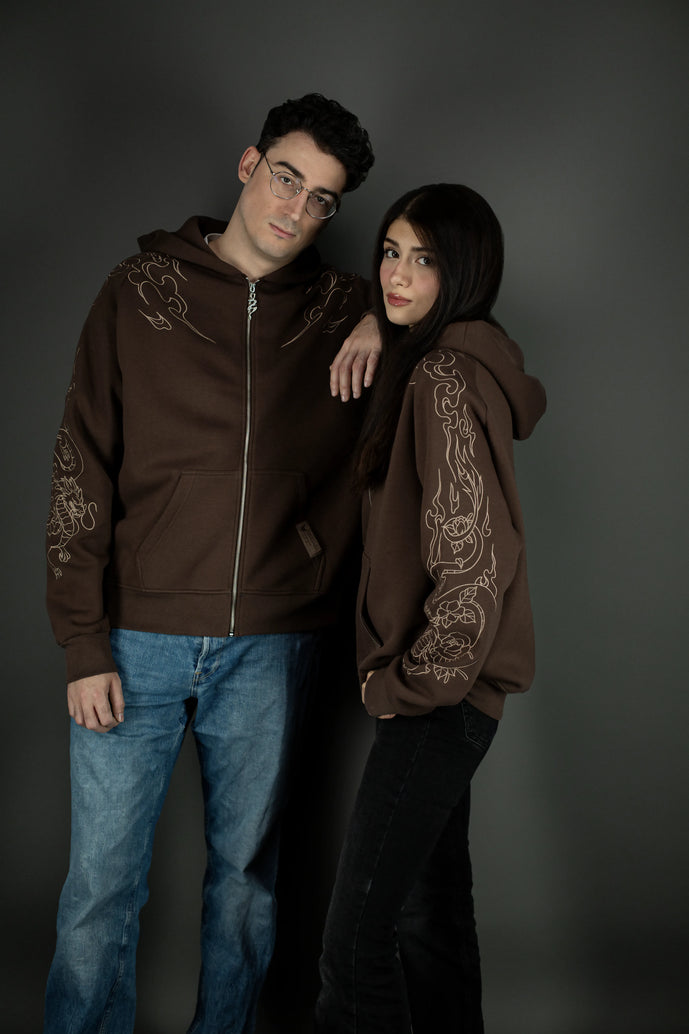 Zwei Personen tragen einen braunen Zip-Hoodie mit beigem Drachenmotiv auf den Ärmeln. Der Hoodie zeigt vorn eine Kängurutasche und Metallreißverschluss. Das Motiv läuft von der Schulter über den Arm und kombiniert Flammen- mit Blumenornamenten.