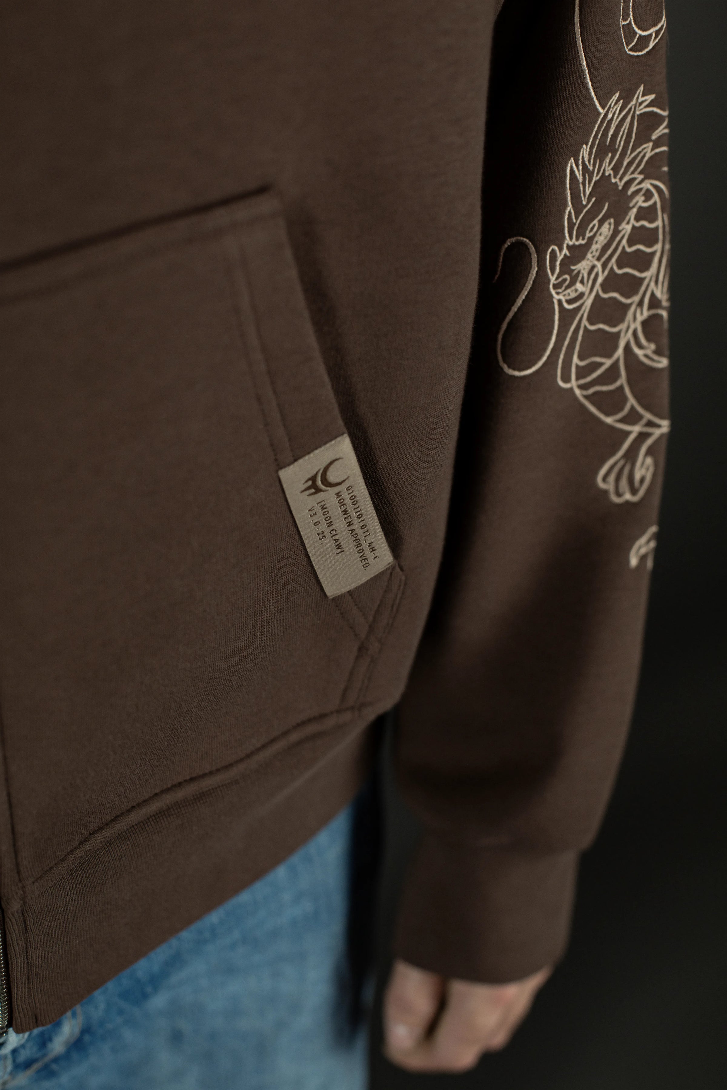 Detailaufnahme der vorderen Kängurutasche des Hoodies. Neben der Tasche ist ein gewebtes Label mit Moonclaw-Logo und Serienangabe „COL. 3.0“ zu sehen.