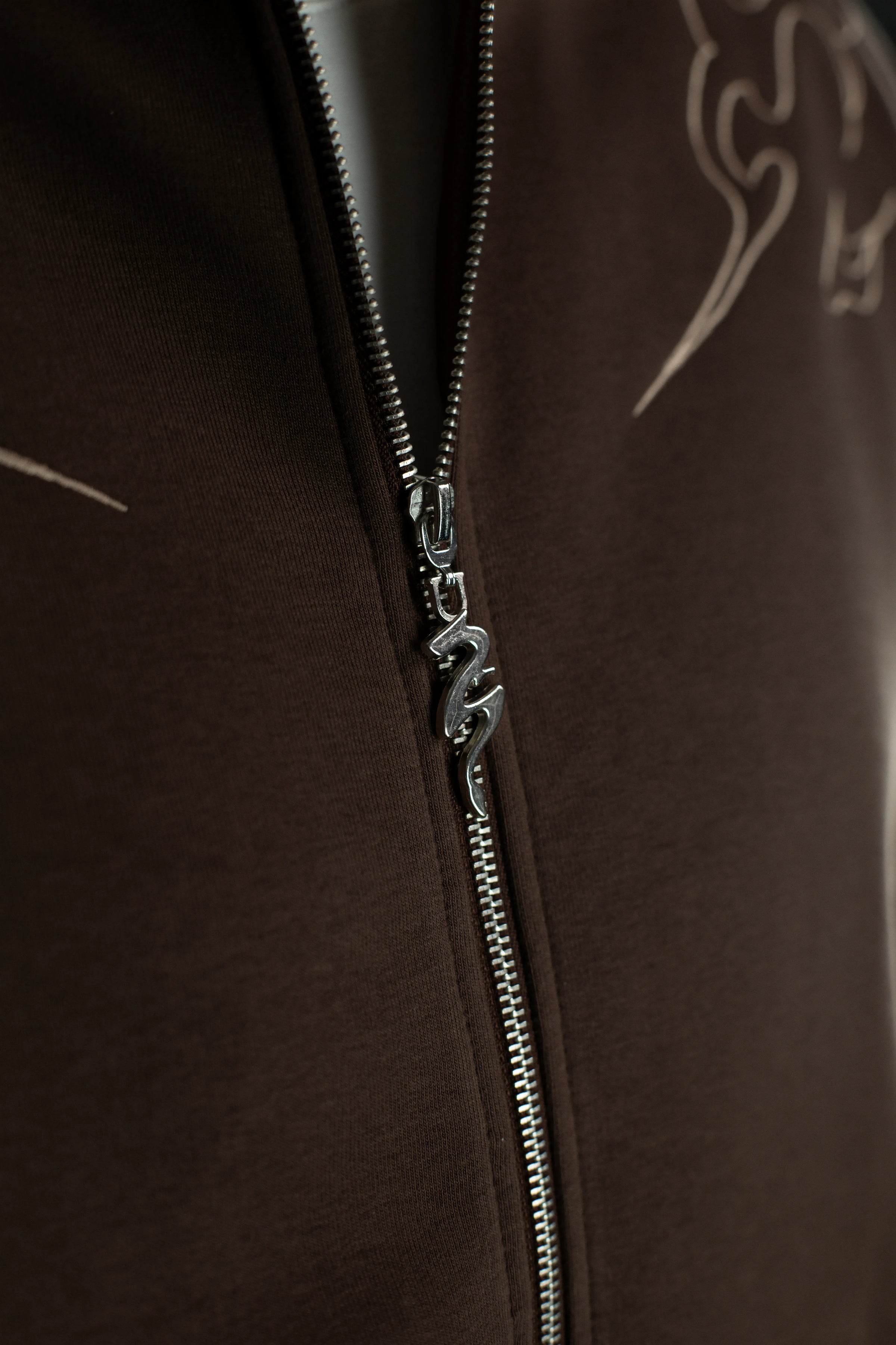 Detail des silbernen Reißverschlusses des Hoodies, dessen Zipper in Form einer geschwungenen Drachenfigur gestaltet ist. Der Stoff ist in dichter, glatter Struktur sichtbar.